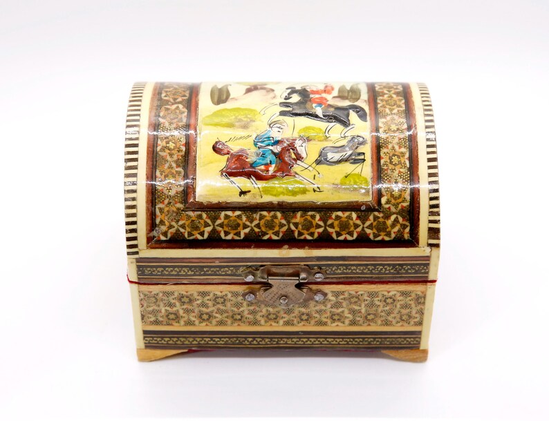 May include: A small, arched wooden chest with a hinged lid. The chest is adorned with intricate mosaic patterns and a painted scene of figures on horseback. The color palette includes beige, brown, red, and gold, with a metal clasp closure.