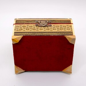 May include: A small, rectangular box with a hinged lid. The box features a red velvet body, wooden corners, and a decorative top with intricate patterns in shades of beige and brown. A metal clasp secures the lid.