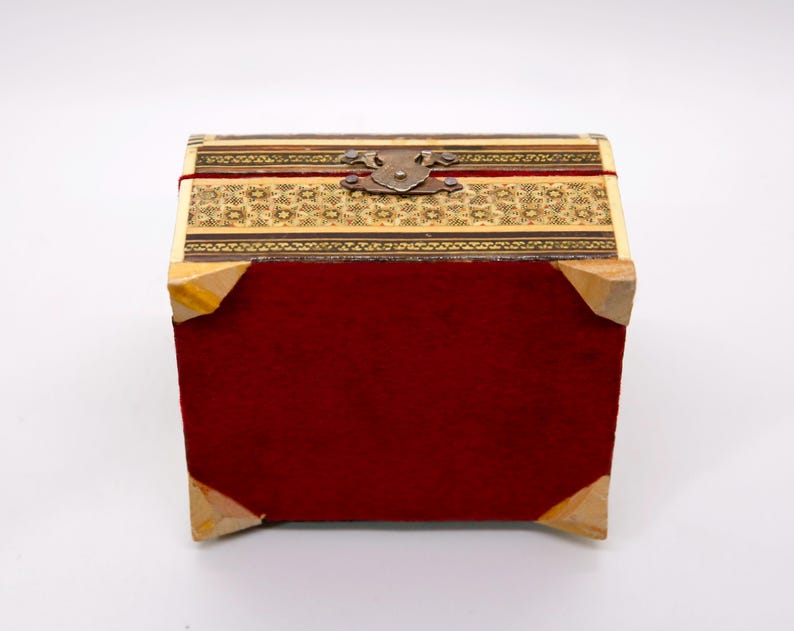 May include: A small, rectangular box with a hinged lid. The box features a red velvet body, wooden corners, and a decorative top with intricate patterns in shades of beige and brown. A metal clasp secures the lid.