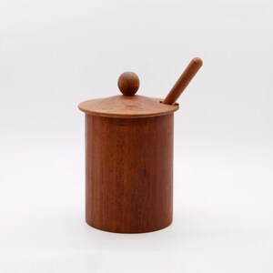 Puede incluir: Un recipiente cilíndrico de madera con tapa y una cuchara pequeña. El recipiente es de madera marrón. La tapa tiene un pequeño pomo en la parte superior y la cuchara descansa en el lateral. La madera tiene un tono marrón cálido.