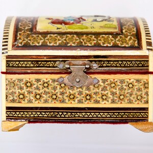 May include: A decorative, arched-top wooden chest with intricate inlaid patterns and a metal clasp. The chest features a small painting on the lid depicting figures on horseback. The exterior is adorned with geometric designs in various colors.