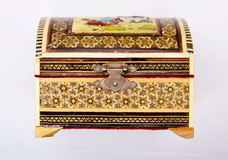 May include: A decorative, arched-top wooden chest with intricate inlaid patterns and a metal clasp. The chest features a small painting on the lid depicting figures on horseback. The exterior is adorned with geometric designs in various colors.