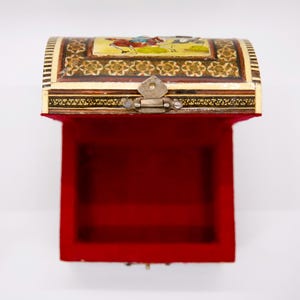 May include: An open, ornate chest with a red velvet interior. The exterior is cream-colored with intricate gold and brown detailing. The lid features a painted scene with figures. A metal clasp secures the lid.