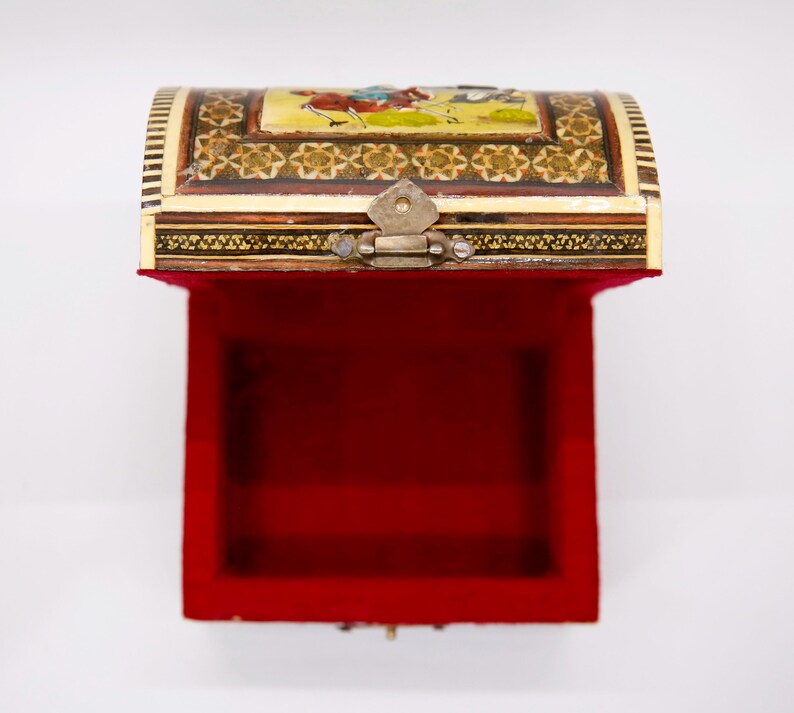 May include: An open, ornate chest with a red velvet interior. The exterior is cream-colored with intricate gold and brown detailing. The lid features a painted scene with figures. A metal clasp secures the lid.
