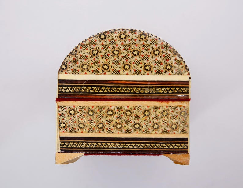 May include: A decorative wooden box with a domed lid. The box features intricate inlaid patterns in shades of beige, brown, and red. The design includes floral and geometric motifs, with a dark border and a red accent.