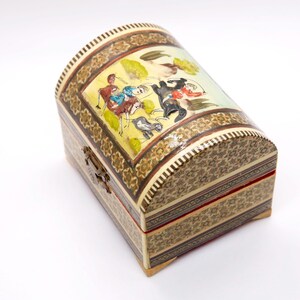 May include: A decorative wooden chest with a curved lid. The chest features intricate mosaic patterns and a painted scene on the lid depicting figures on horseback. The color palette includes beige, brown, yellow, and red accents. A metal clasp secures the front.