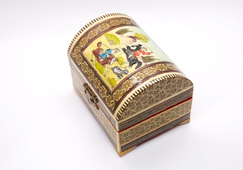 May include: A decorative wooden chest with a curved lid. The chest features intricate mosaic patterns and a painted scene on the lid depicting figures on horseback. The color palette includes beige, brown, yellow, and red accents. A metal clasp secures the front.