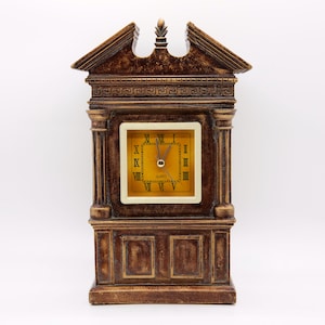 May include: A brown decorative quartz clock with Roman numerals on a gold face. The clock has a rectangular shape with a decorative top and base. The clock is designed to resemble a building facade.