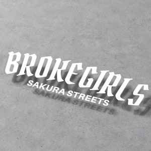 Könnte beinhalten: Weißer Aufkleber mit dem Text "BROKEGIRLS" in einer stilisierten Schriftart. Unter dem Text befindet sich der Satz "SAKURA STREETS" in einer kleineren Schriftart.