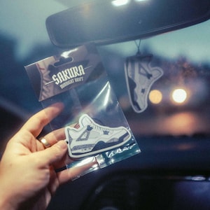 May include: A car interior shot showing a hand holding a clear plastic packet with a sneaker-shaped air freshener. The packet reads "SAKURA MIDNIGHT DRIFT". Another sneaker air freshener hangs from the rearview mirror.