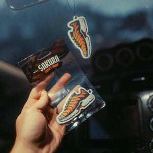 May include: Car air fresheners in the shape of orange and white sneakers. One is hanging from a rearview mirror that says "Ghost Boy." Another is in a clear plastic package with the text "Sakura Sunset Sauce."