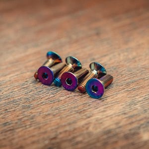 Op de afbeelding: Vijf iriserende schroeven met een platte kop en een zeskant-aandrijving. De schroeven zijn in een rij op een houten oppervlak gerangschikt.