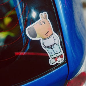 Puede incluir: Un coche azul con una pegatina de un perro de dibujos animados en la ventana trasera. El perro lleva una camisa blanca y pantalones vaqueros azules y tiene un zapato rojo y blanco.