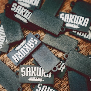 Peut inclure: Un ensemble de porte-clés noirs avec du texte blanc. Les porte-clés présentent les mots "Sakura" et "Vanquish".