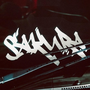 Op de afbeelding: Witte tekst in graffiti-stijl op een zwarte ondergrond. De tekst luidt "SAKURA" in een gestileerd lettertype. De afbeelding is vanuit een hoek genomen, met ruitenwissers zichtbaar.