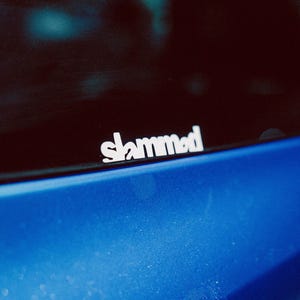 Puede incluir: Primer plano de una ventana de coche con una pegatina blanca con la palabra "slammed". La pegatina está colocada encima de una sección de pintura azul brillante. La imagen tiene una estética ligeramente granulada y vintage.