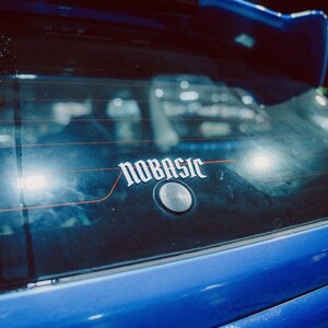May include: A close-up of the rear of a blue car with a spoiler. The car's rear window has a white sticker with the word "NOBASIC" in a stylized font. A silver button is below the sticker.