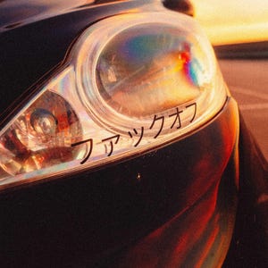 May include: A close-up of a car's headlight with a sticker that says "ワッツアップ" in Japanese.