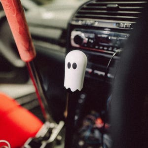 Op de afbeelding: Een witte spookvormige versnellingspookknop met zwarte ogen is het middelpunt van deze afbeelding. De knop is bevestigd aan een zwarte versnellingspook, geplaatst in het interieur van een auto. Een rode handgreep is zichtbaar aan de linkerkant. Het dashboard en de radio van de auto zijn op de achtergrond.