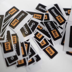 Puede incluir: Un montón de pegatinas rectangulares con la inscripción "Good Boy". El texto es blanco sobre fondo negro, con un borde naranja. Las pegatinas están dispersas sobre una superficie blanca.