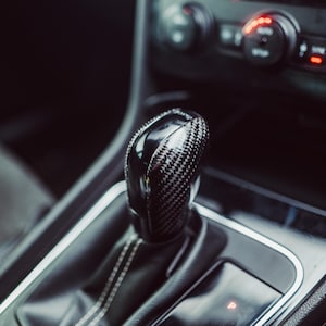 Könnte beinhalten: Nahaufnahme des Schalthebels eines Autos. Der Schaltknauf ist schwarz mit einem Karbonfasermuster. Die Ledermanschette um den Schalthebel hat weiße Nähte. Die Gangwahltaste zeigt die Buchstaben P, R, N, D/S und Plus- und Minuszeichen.