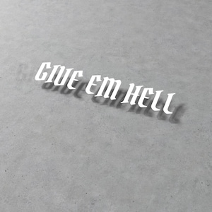 Op de afbeelding: Witte tekst "GIVE EM HELL" in gotisch lettertype op een grijze betonnen ondergrond. De tekst werpt een schaduw, wat diepte creëert. De afbeelding heeft een minimalistische uitstraling.