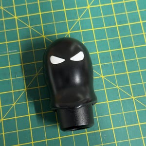 May include: A black shift knob with a glossy finish. The knob has two white, angular eye shapes. The base is cylindrical. The background is a green cutting mat with yellow grid lines.