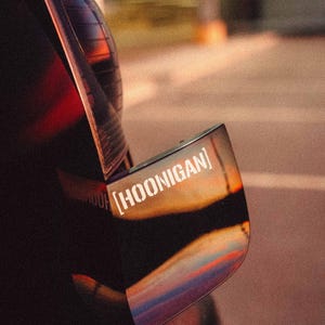 Puede incluir: Primer plano de un coche negro con un espejo lateral reflectante. El espejo tiene una pegatina blanca con la palabra "[HOONIGAN]" impresa en él.