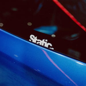Może przedstawiać: Zbliżenie niebieskiego samochodu z czarną szybą. Na szybie naklejona jest biała naklejka "Static". Na szybie widoczny jest czerwony pasek. Lakier samochodu ma błyszczące wykończenie.
