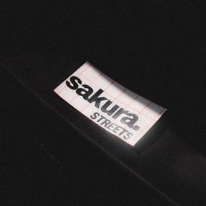 Peut inclure: Un autocollant blanc avec du texte noir qui dit "sakura. streets" sur un fond noir.