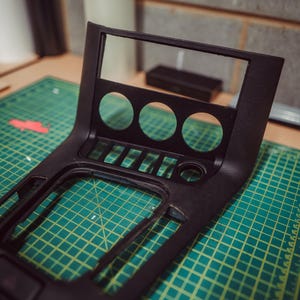 May include: A black car dashboard centre console with three circular openings, several rectangular slots, and a rectangular opening. The console is on a green cutting mat with a grid pattern.