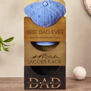 May include: A three-tiered wooden hat rack with the words "Best Dad Ever", "Jacob's Rack", and "Dad" engraved on each tier. The top tier has a blue baseball cap on it. The bottom tier has the names "Melinda, David, Herbert" engraved on it.