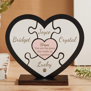 May include: A black heart-shaped wooden plaque with a jigsaw puzzle design. The puzzle pieces are labeled with names: "Bridget", "Joyce", "Crystal", and "Lucky". A pink heart in the center reads "Mom You are the piece that holds us together".