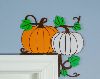 Puede incluir: Un tope de puerta de metal marrón en forma de calabaza con dos calabazas, una naranja y una blanca, con hojas verdes.