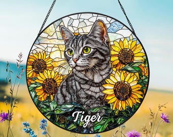 May include: A circular stained glass suncatcher featuring a grey tabby cat with green eyes surrounded by yellow sunflowers. The name Tiger is written in white script at the bottom. The piece is framed in black metal and hangs from a black chain.