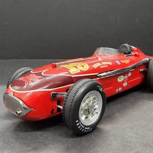 May include: A red and gold 1950s era race car model with a black driver's helmet and a red horse logo on the side. The car has black wheels and a chrome bumper.