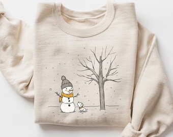 Puede incluir: Sudadera beige con una ilustración de una escena invernal. El diseño muestra un muñeco de nieve con gorro gris y bufanda amarilla, un perro pequeño y un árbol sin hojas. La ilustración es en blanco y negro con toques de color. La sudadera está doblada.