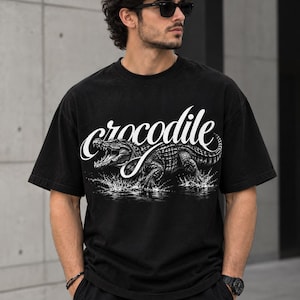 Może przedstawiać: Czarny t-shirt z krótkim rękawem z białym nadrukiem graficznym przedstawiającym krokodyla wyłaniającego się z wody. Słowo crocodile jest napisane stylizowaną białą kursywą nad ilustracją gada na przedniej części odzieży.