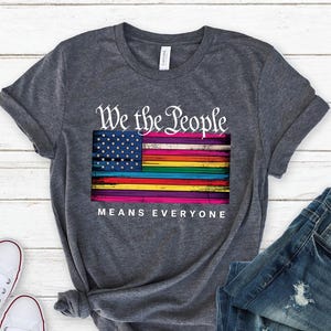 May include: Dark gray t-shirt with the text "We the People Means Everyone" and a colorful flag design. The flag features a blue field of stars and rainbow stripes. White sneakers and blue jeans are also in the image.
