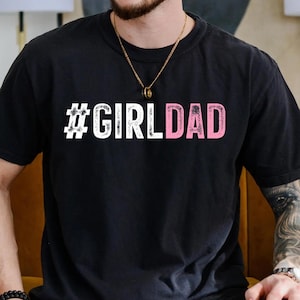 May include: Black t-shirt with a white hashtag and the text "#GIRLDAD" in pink.