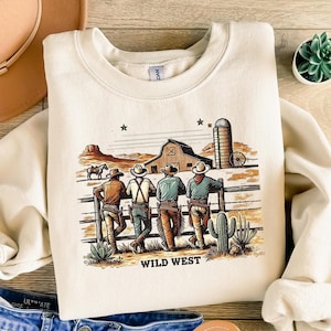 Könnte beinhalten: Ein cremefarbener Pullover mit einem Grafikdruck von vier Cowboys, die vor einem Holzzaun stehen. Die Grafik enthält auch eine Scheune, einen Silo und Kakteen. Der Text "WILD WEST" ist unterhalb der Grafik gedruckt.