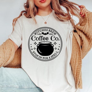 以下が含まれることがあります： 「Witches Brew Coffee Co. Stop in for a spell.」と書いてある黒と白のグラフィックデザインが施された白いTシャツ。グラフィックには、釜、コウモリ、星が描かれています。