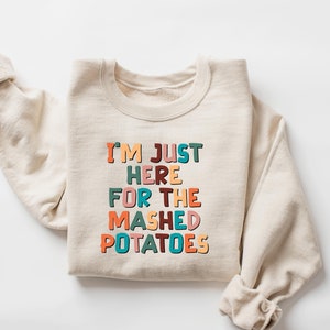 Puede incluir: Una sudadera beige con el texto "I'm Just Here For The Mashed Potatoes" impreso en letras de colores.