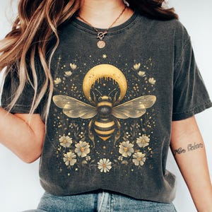 May include: Dark gray t-shirt featuring a detailed illustration of a bee with a crescent moon and floral elements. The bee is in the center, with a yellow moon above and flowers below. The design has gold and white accents.