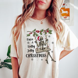 May include: A cream colored t-shirt with a skeleton on a swing decorated with Christmas lights and holly. The text on the shirt reads "Have a Holly Jolly Christmas".