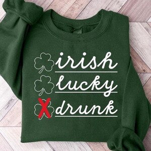 以下が含まれることがあります： 「Irish lucky drunk」と白文字で書かれた緑色のスウェットシャツ。drunkの文字の上に赤いXが書かれている。