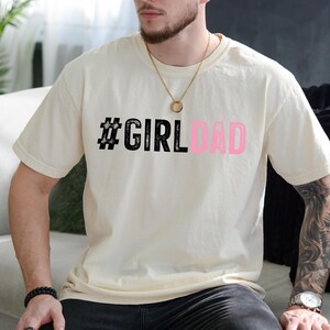 May include: A white t-shirt with the text "#GIRLDAD" printed in black and pink. The text is in a bold, stylized font.
