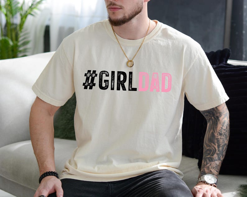 May include: A white t-shirt with the text "#GIRLDAD" printed in black and pink. The text is in a bold, stylized font.
