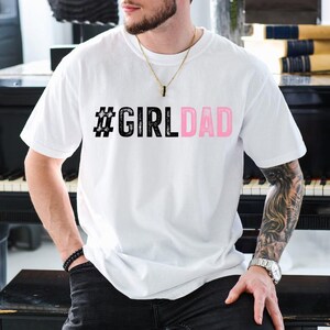 May include: White t-shirt with a black hashtag and the text "#GIRLDAD" in pink letters.