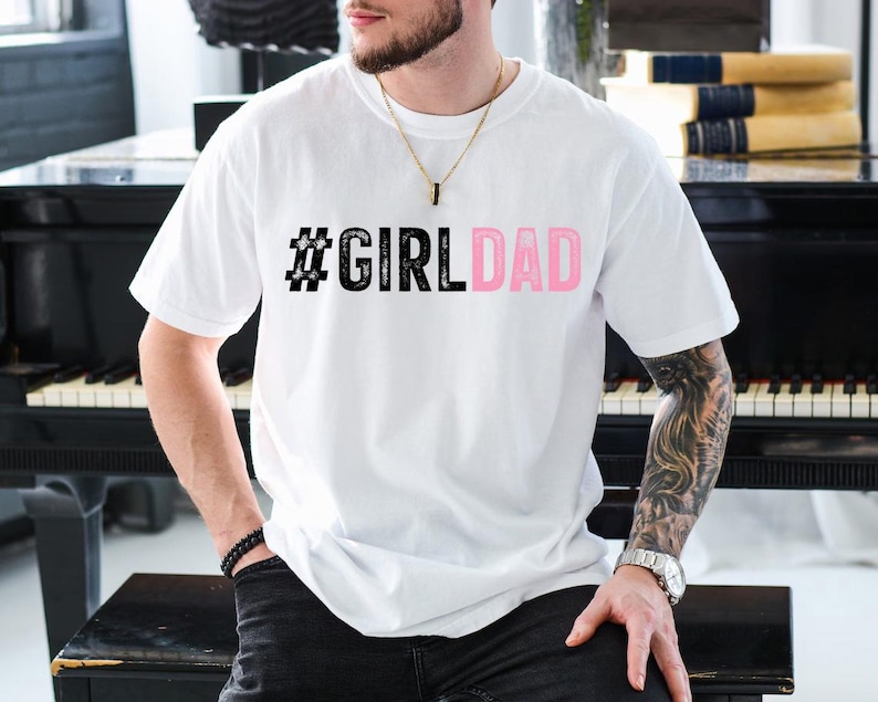May include: White t-shirt with a black hashtag and the text "#GIRLDAD" in pink letters.